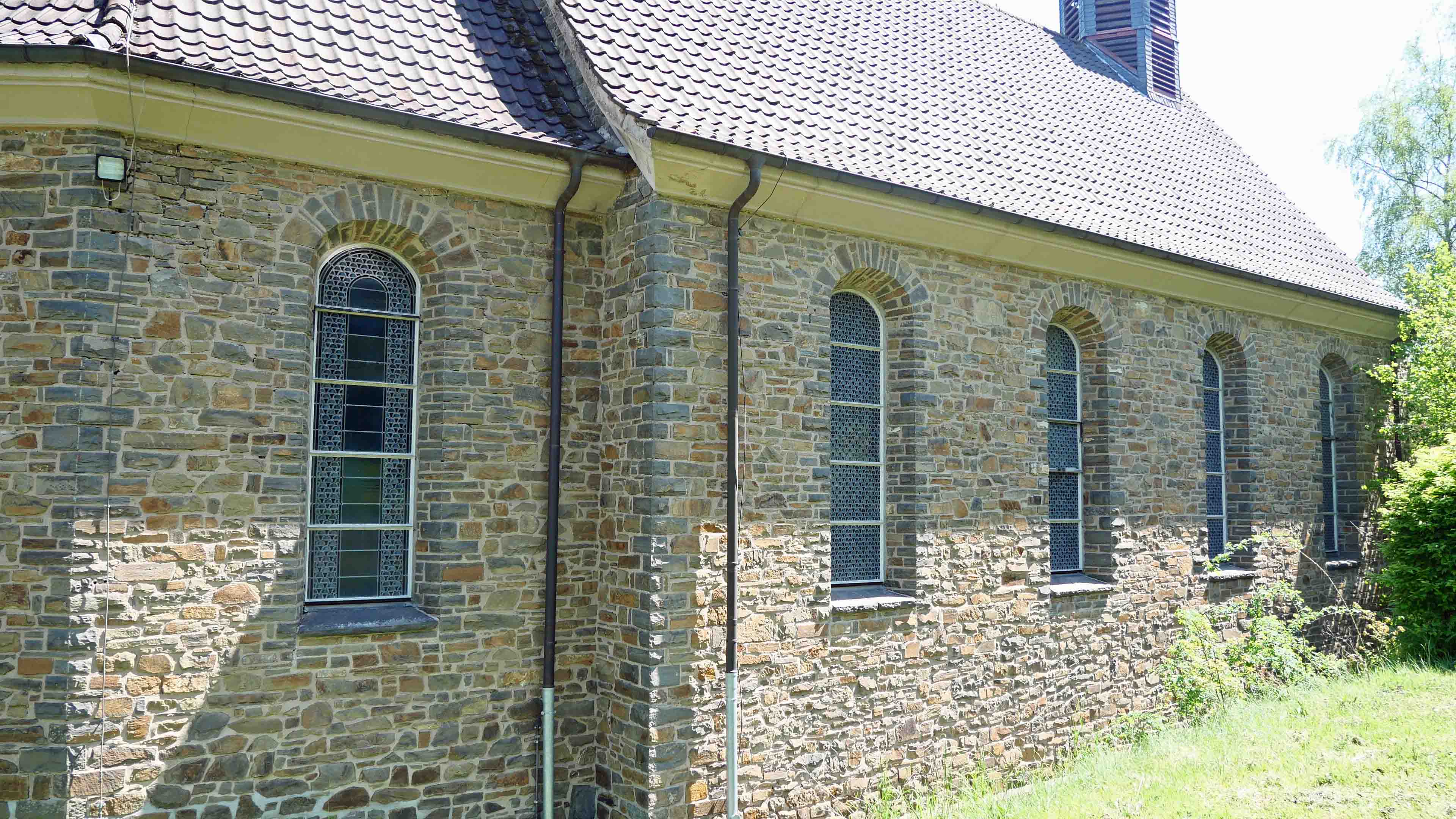Morsbach-Ellingen: Stilvolle, kleine Bruchsteinkirche, Westseite mit großen Mosaik-Fenstern